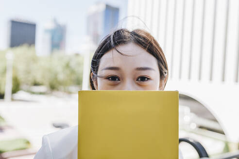 Female entrepreneur covering face with book while standing against financial district - MRRF00433