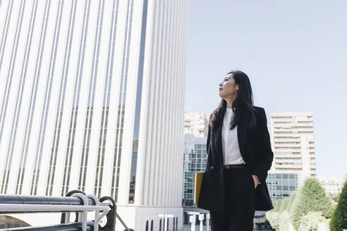 Businesswoman with hands in pockets wearing blazer jacket while standing against financial district - MRRF00434