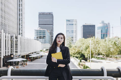 Confident businesswoman with book standing against downtown district in city - MRRF00435