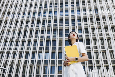 Businesswoman holding book while standing against downtown district in city - MRRF00438