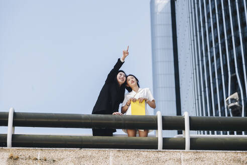 Smiling businesswoman pointing female coworker at downtown district in city against clear sky - MRRF00439