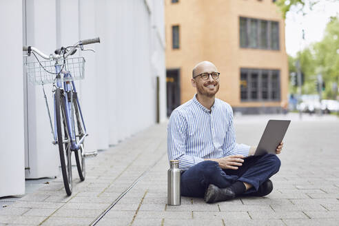 Lächelnder erwachsener Mann, der einen Laptop benutzt und auf der Straße sitzt - MCF01493