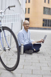 Mittlerer Erwachsener, der an einem Laptop arbeitet, während er auf der Straße sitzt - MCF01494