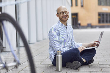 Glücklicher mittlerer Erwachsener, der an einem Laptop arbeitet, während er auf der Straße sitzt - MCF01495