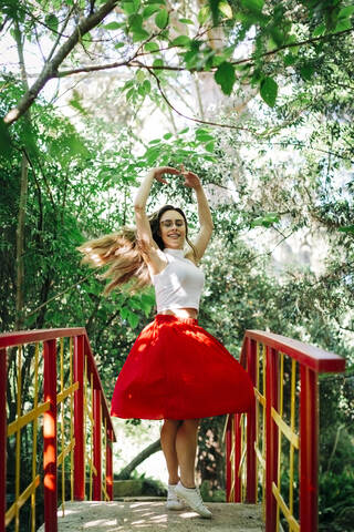 Smiling woman with arms raised practicing ballet on footbridge against trees in park stock photo