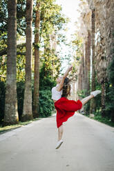 Young woman jumping while dancing on road amidst trees in park - DCRF00835