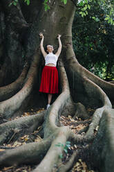 Junge Frau mit erhobenen Armen an einen Baumstamm gelehnt im Park - DCRF00837