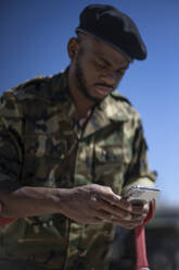 Militäroffizier mit Krücken, der an einem sonnigen Tag eine Textnachricht auf seinem Smartphone schreibt - OCMF01690
