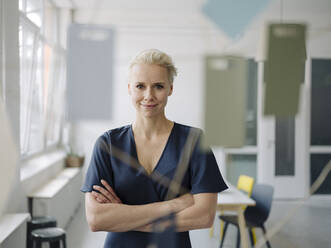 Confident businesswoman with arms crossed standing in office seen through window - KNSF08549