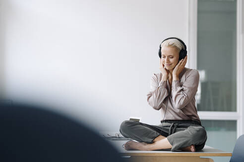 Lächelnde Geschäftsfrau mit geschlossenen Augen, die über Kopfhörer Musik hört, während sie am Schreibtisch sitzt - KNSF08558