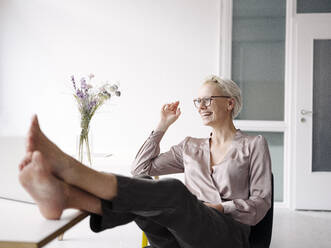 Smiling businesswoman with feet on desk relaxing in loft office - KNSF08563