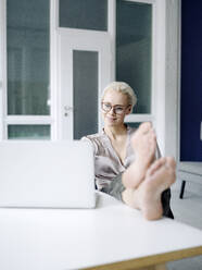 Geschäftsfrau mit Füßen auf dem Schreibtisch, die einen Laptop in einem Loftbüro benutzt - KNSF08564