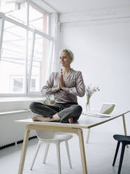 Geschäftsfrau meditiert auf dem Schreibtisch in einem Loftbüro sitzend - KNSF08568