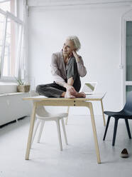 Smiling businesswoman contemplating while sitting on desk in office - KNSF08571