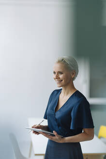 Smiling female entrepreneur with digital tablet looking away while standing in office - KNSF08577