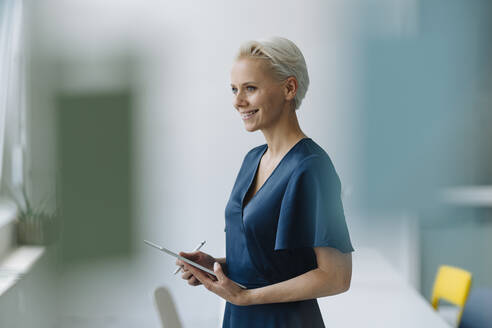 Smiling businesswoman with digital tablet looking away while standing in office - KNSF08578