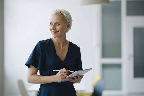 Smiling female entrepreneur holding digital tablet contemplating while standing in office - KNSF08585