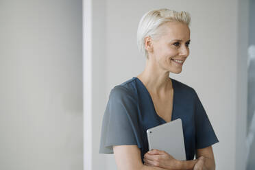 Lächelnde Geschäftsfrau, die ein digitales Tablet hält und wegschaut, während sie im Büro an der Wand steht - KNSF08589