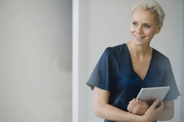 Lächelnde Unternehmerin mit digitalem Tablet, die wegschaut, während sie im Büro an der Wand steht - KNSF08590