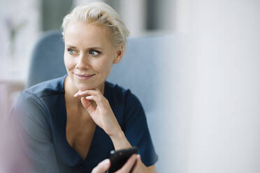 Close-up of thoughtful businesswoman holding smart phone while sitting in office - KNSF08603