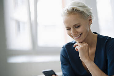 Close-up of smiling female entrepreneur with hand on chin using smart phone in office - KNSF08604