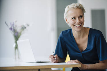 Cheerful businesswoman with laptop on desk looking away while sitting in office - KNSF08605