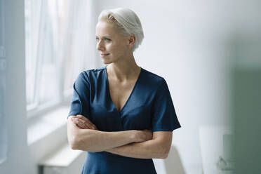 Thoughtful female entrepreneur with arms crossed looking away while standing in office - KNSF08614