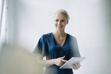 Female professional using digital tablet looking away while standing against wall in office - KNSF08621