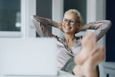 Thoughtful businesswoman with hands behind head relaxing at desk in office - KNSF08624