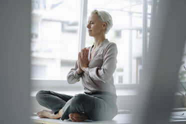 Businesswoman with eyes closed meditating while sitting against window in loft office - KNSF08627