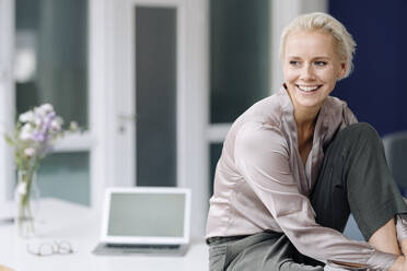 Smiling businesswoman contemplating while sitting on desk in loft office - KNSF08633