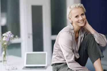 Smiling businesswoman with laptop relaxing on desk in loft office - KNSF08634