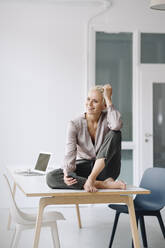 Thoughtful female entrepreneur holding smart phone while sitting on desk against wall in office - KNSF08641