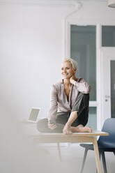 Thoughtful female entrepreneur with smart phone sitting on desk against wall in office - KNSF08642