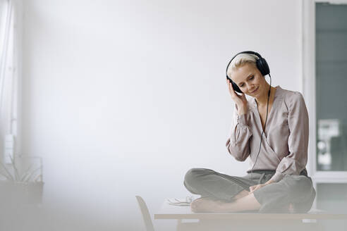 Weibliche Fachkraft hört Musik über Kopfhörer, während sie auf dem Schreibtisch an der Wand im Büro sitzt - KNSF08651