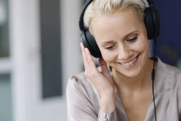 Close-up of smiling businesswoman listening music over headphones looking down in office - KNSF08654