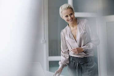 Smiling businesswoman looking away while using laptop against wall in office - KNSF08665