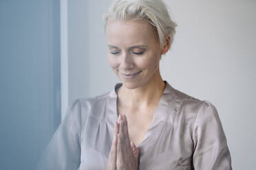 Close-up of smiling businesswoman with eyes closed meditating against wall in office - KNSF08677