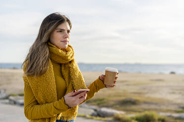 Thoughtful woman with mobile phone and coffee cup looking away against sky - AFVF07245