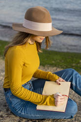 Junge Frau mit Sonnenhut schreibt in Tagebuch, während sie auf dem Boden sitzt - AFVF07252