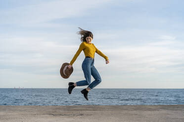 Junge Frau mit Sonnenhut springt beim Genießen gegen das Meer - AFVF07256