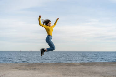 Young woman with hand raised jumping while enjoying against sea - AFVF07257