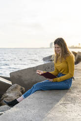 Young woman with diary sitting by sea against sky - AFVF07265