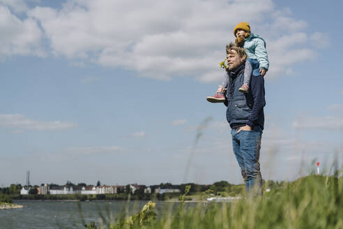 Vater steht mit den Händen in den Taschen und trägt seine Tochter auf der Schulter in der Nähe des Rheins - GUSF04440