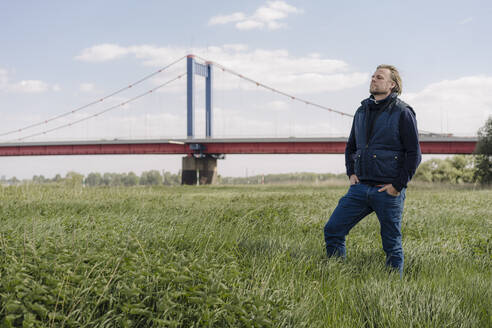 Älterer Mann im Gras stehend mit Händen in den Taschen vor einer Brücke - GUSF04442