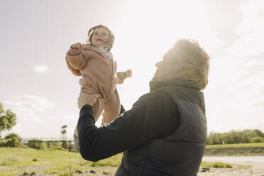 Vater, der seine kleine Tochter abholt, während er an einem sonnigen Tag am Flussufer steht - GUSF04471