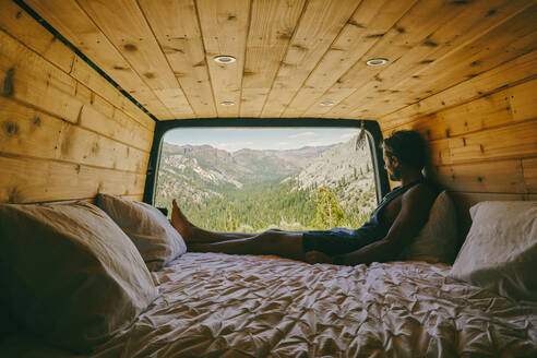 Junger Mann sitzt auf dem Bett eines Wohnmobils und betrachtet die Aussicht auf den Yosemite - CAVF88877