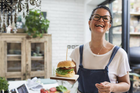 Portrait laughing woman holding cheeseburger on cutting board - CAIF29534