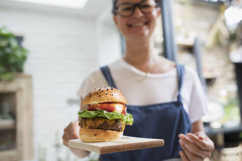 Portrait smiling woman with cheeseburger on cutting board - CAIF29618
