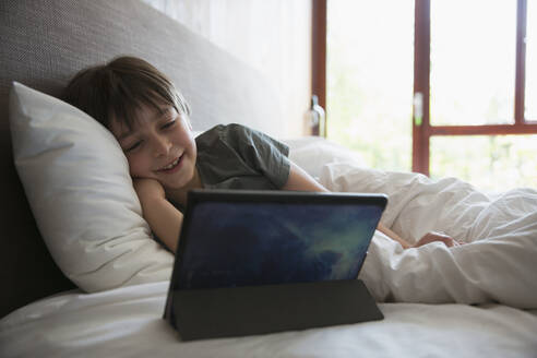 Lächelnder Junge mit digitalem Tablet im Bett - CAIF29636